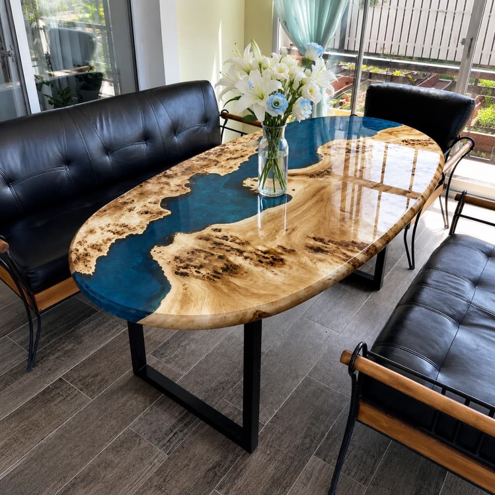 Wide interior perspective of casual residential café dining zone with grey wood-look tile flooring, sheer mint window curtains and full leather seating framing organic oval maple blue river epoxy table