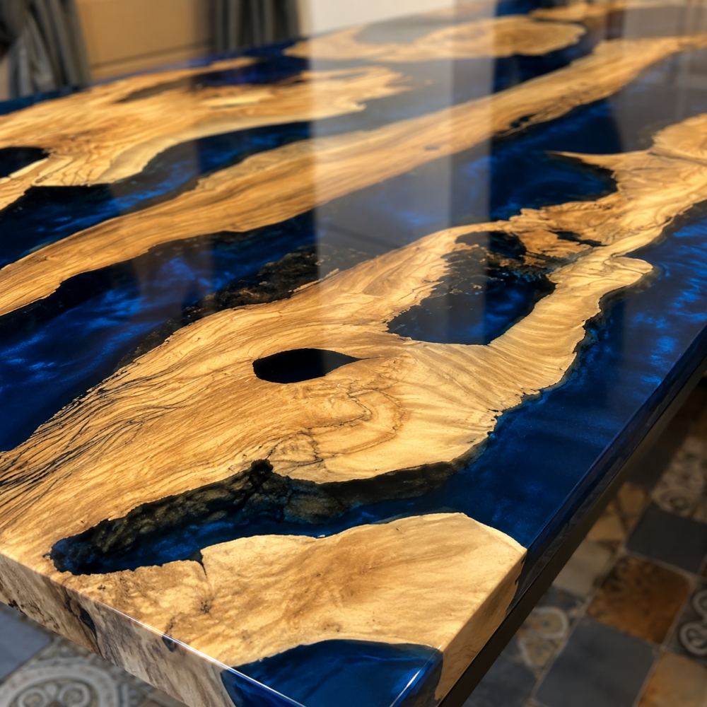 Diagonal overhead cabin interior shot of multi-piece live edge olive wood dining table wrapped in glossy deep sapphire blue epoxy river, surrounded by dark gray padded upholstered dining chairs, light horizontal wood plank walls and heavy charcoal draped window curtains